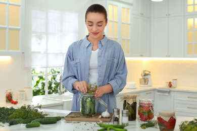 Woman putting dill into pickling jar at table in kitchen