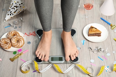 Woman using scale surrounded by food and alcohol after party on floor. Overweight problem