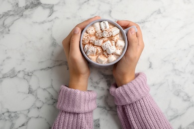 Woman in sweater holding cup of tasty chocolate with milk and marshmallows on marble table, top view