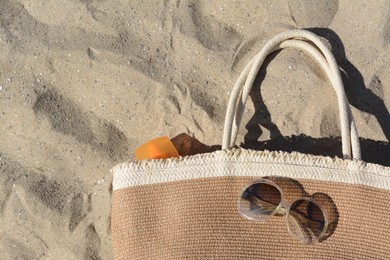 Photo of Straw bag with sun protection product and sunglasses on sand, top view. Beach accessories