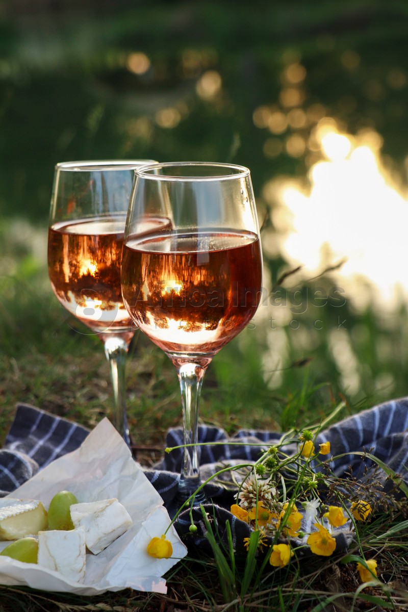 Glasses of delicious rose wine, cheese and grapes on picnic blanket near lake
