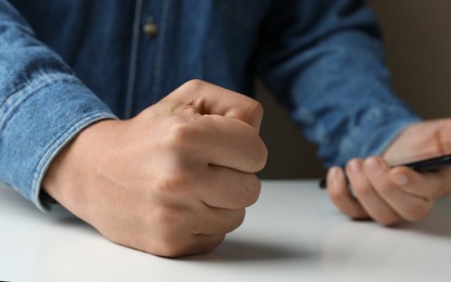 Photo of Man with clenched fist holding mobile phone at table, closeup. Restraining anger