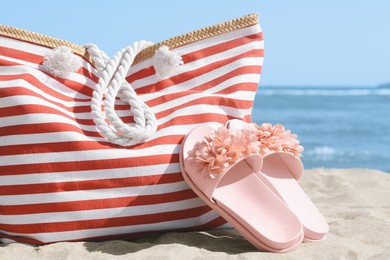 Stylish striped bag with slippers on sandy beach near sea, closeup