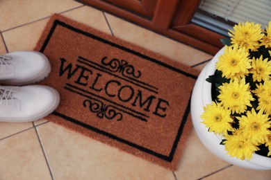 Door mat with word Welcome, stylish boots and beautiful flowers on floor, above view