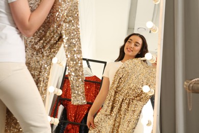 Photo of Beautiful young woman with golden sequin dress near mirror in boutique. Choosing party clothes