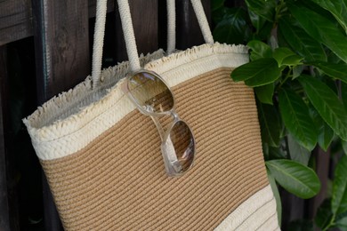 Stylish bag with sunglasses hanging on wooden fence outdoors, closeup. Beach accessories