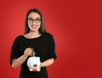 Young woman putting money into piggy bank on crimson background. Space for text