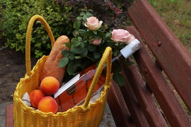 Photo of Yellow wicker bag with roses, peaches, baguette and wine on wooden bench outside. Space for text
