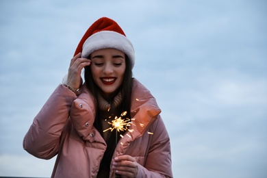 Woman in Santa hat and warm clothes holding burning sparkler outdoors