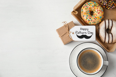 Tag with phrase HAPPY FATHER'S DAY, donuts, coffee and gift on white wooden table, flat lay. Space for text