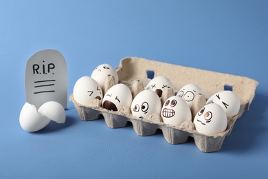 Eggs with drawn faces and paper grave near broken shell on blue background. April fool's day