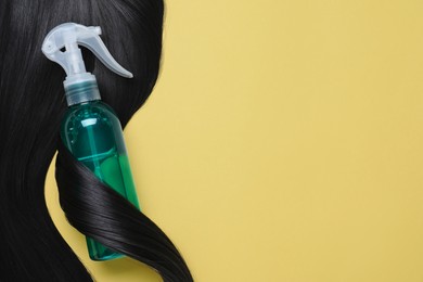 Photo of Spray bottle with thermal protection wrapped in lock of black hair on yellow background, top view. Space for text