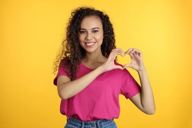 Happy young African-American woman making heart with hands on yellow background