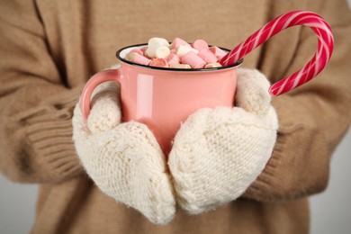 Woman in knitted mittens holding cup of delicious hot chocolate with marshmallows and candy cane, closeup