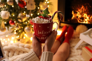 Woman holding cup of sweet drink near fireplace at home, closeup. Cozy winter holidays atmosphere