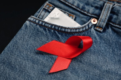 Condom and red ribbon in pocket of jeans on black background, closeup. AIDS disease awareness