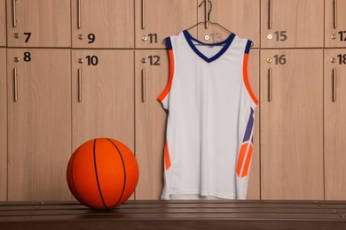Photo of Orange basketball ball on wooden bench and hanger with uniform in locker room