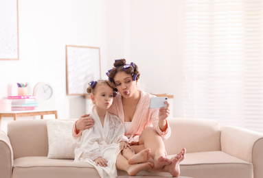 Happy mother and daughter with curlers and mobile phone on sofa at home