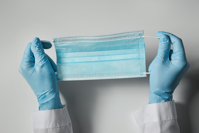 Doctor in latex gloves holding disposable face mask on light background, closeup. Protective measures during coronavirus quarantine