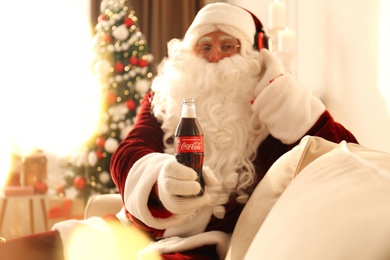 MYKOLAIV, UKRAINE - JANUARY 18, 2021: Santa Claus listening to music with headphones at home, focus on Coca-Cola bottle in his hand