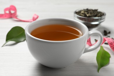 Cup of herbal diet tea, measuring tape and green leaves on white wooden table, closeup. Weight loss concept