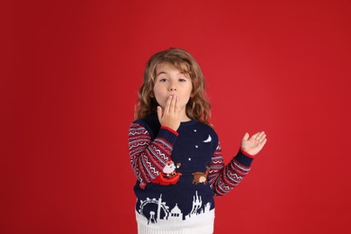 Little girl in Christmas sweater covering her mouth against red background