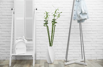 Vase with green bamboo stems, mirror and rack on floor near white brick wall in room. Interior design