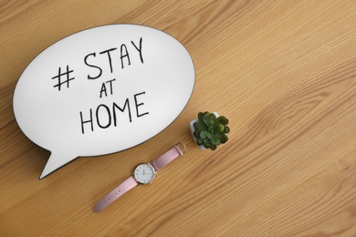 Houseplant, wristwatch and speech bubble with hashtag STAY AT HOME on wooden background, flat lay. Message to promote self-isolation during COVID‑19 pandemic