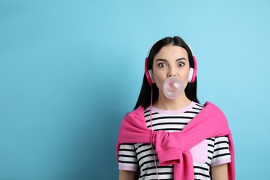 Fashionable young woman with headphones blowing bubblegum on light blue background, space for text