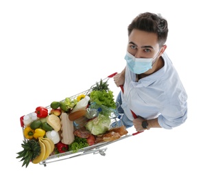 Young man in medical mask with shopping cart full of groceries on white background, above view