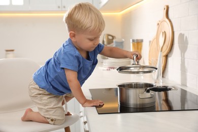 Photo of Curious little boy playing with saucepan on electric stove in kitchen