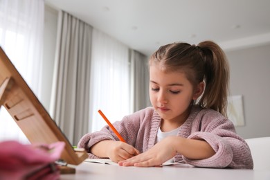 Photo of Adorable little girl doing homework with tablet at table indoors