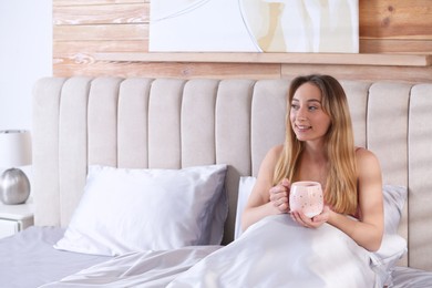 Young woman holding cup of drink on comfortable bed with silky linens, space for text