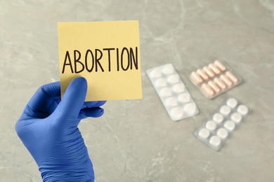 Doctor holding paper card with word ABORTION over light grey marble table with pills, closeup