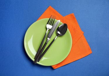 Plate with shiny silver cutlery on blue background, top view