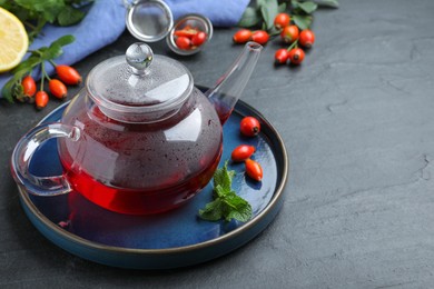 Teapot with aromatic rose hip tea, berries and mint on black table, space for text