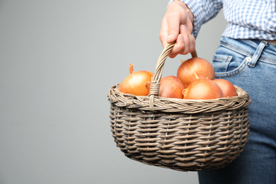 Woman holding wicker basket with raw yellow onion bulbs on grey background, closeup. Space for text