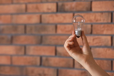 Woman holding light bulb near brick wall indoors, closeup. Space for text