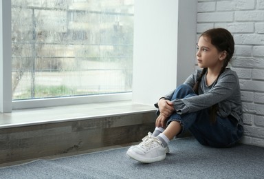 Sad little girl sitting on floor near window indoors, space for text
