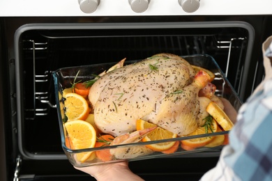 Woman putting raw chicken with orange slices into oven, closeup