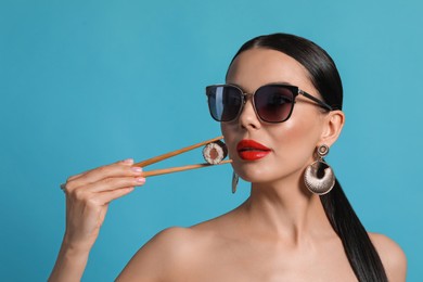 Attractive woman in fashionable sunglasses holding chopsticks with sushi against light blue background