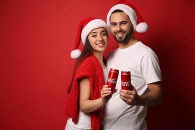 MYKOLAIV, UKRAINE - JANUARY 27, 2021: Young couple in Christmas hats holding cans of Coca-Cola on red background