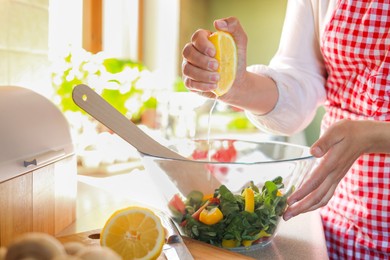 Photo of Woman squeezing fresh lemon over bowl with salad at countertop in kitchen, closeup