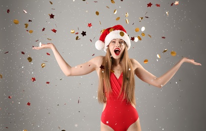 Young beautiful woman in Santa hat and swimsuit with sparkling confetti on grey background. Christmas celebration