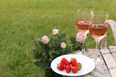 Photo of Picnic basket with glasses of delicious rose wine, strawberries and flowers outdoors, space for text