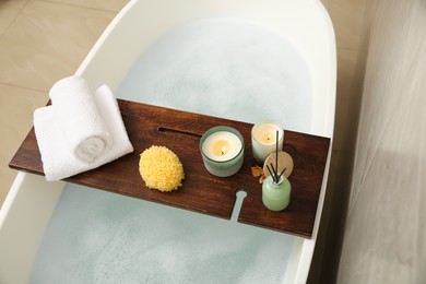 Wooden bath tray with candles, air freshener, towels and sponge on tub indoors, above view
