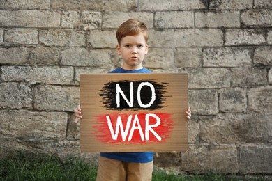 Sad boy holding poster with words No War against brick wall outdoors