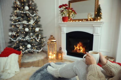 Woman with hot drink resting near fireplace in cozy room decorated for Christmas, closeup