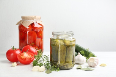 Glass jars of pickled cucumbers and tomatoes on white table