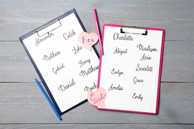 Clipboards with different baby names and felt tip pens on grey wooden table, flat lay
